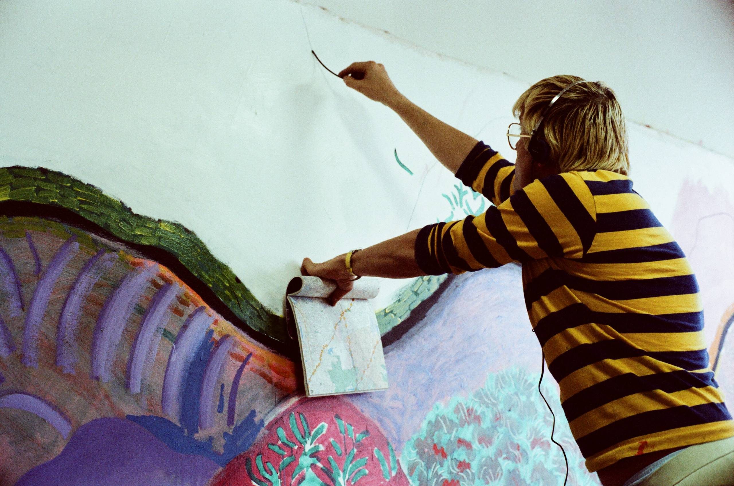 A person wearing glasses and a yellow and black striped shirt paints a colourful mural on a wall, holding a sketchbook for reference in one hand and using the other hand to paint.