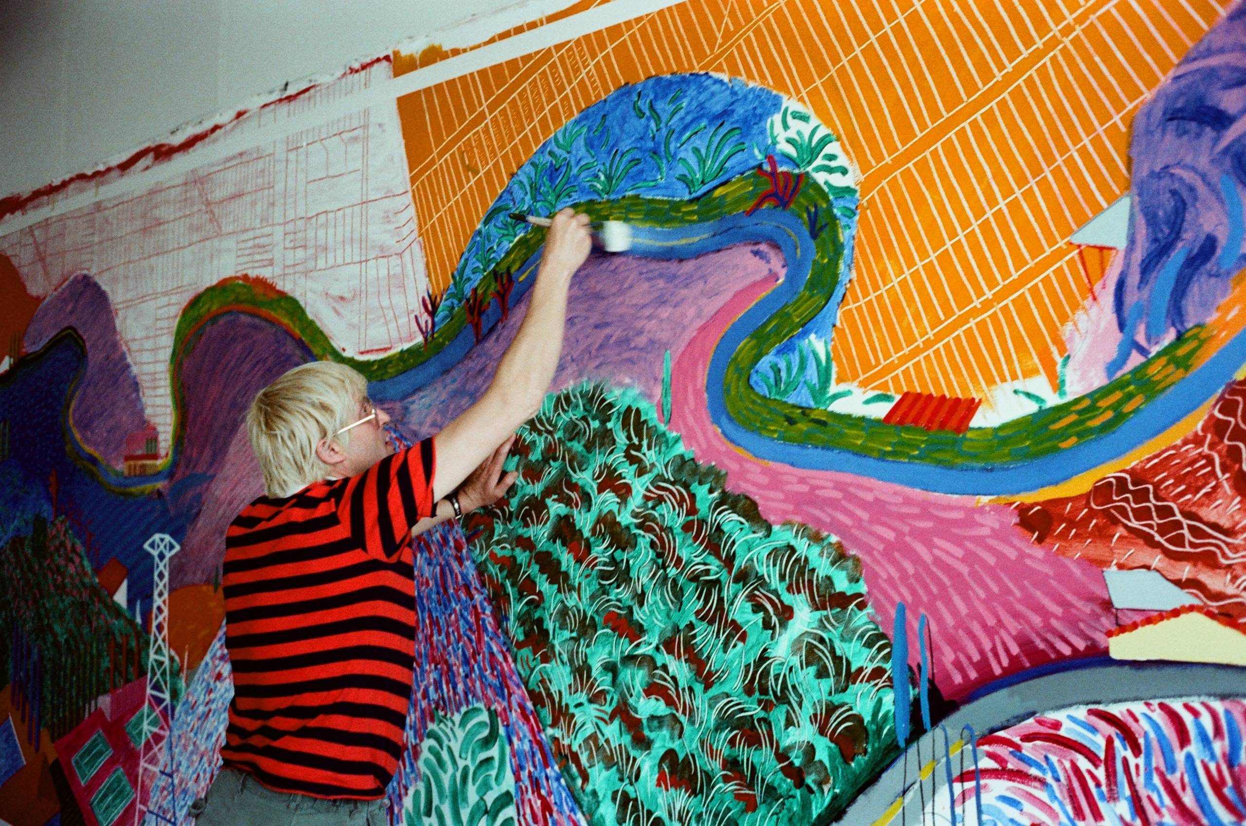 A person with blond hair wearing a red and black striped shirt paints a colourful, abstract mural on a large wall using a wide brush. The mural features bold shapes, plants, and winding river patterns.