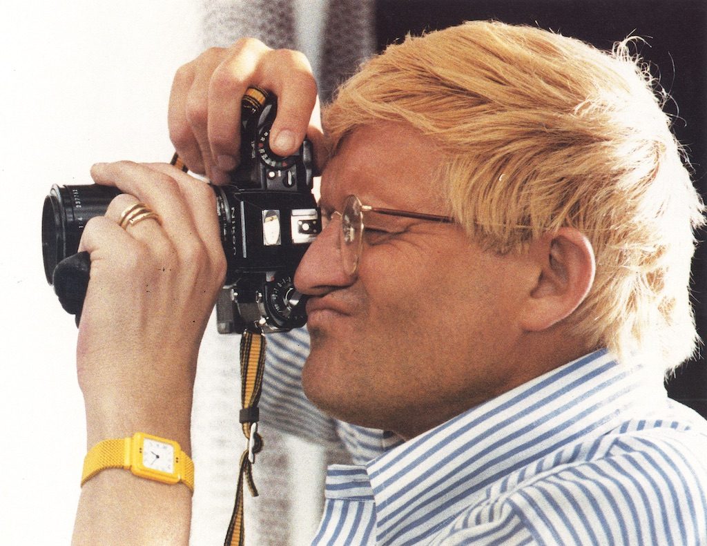 A person with blond hair, wearing glasses, a blue-and-white striped shirt, and a yellow wristwatch, holds a camera up to their face and looks through the viewfinder, preparing to take a photograph.
