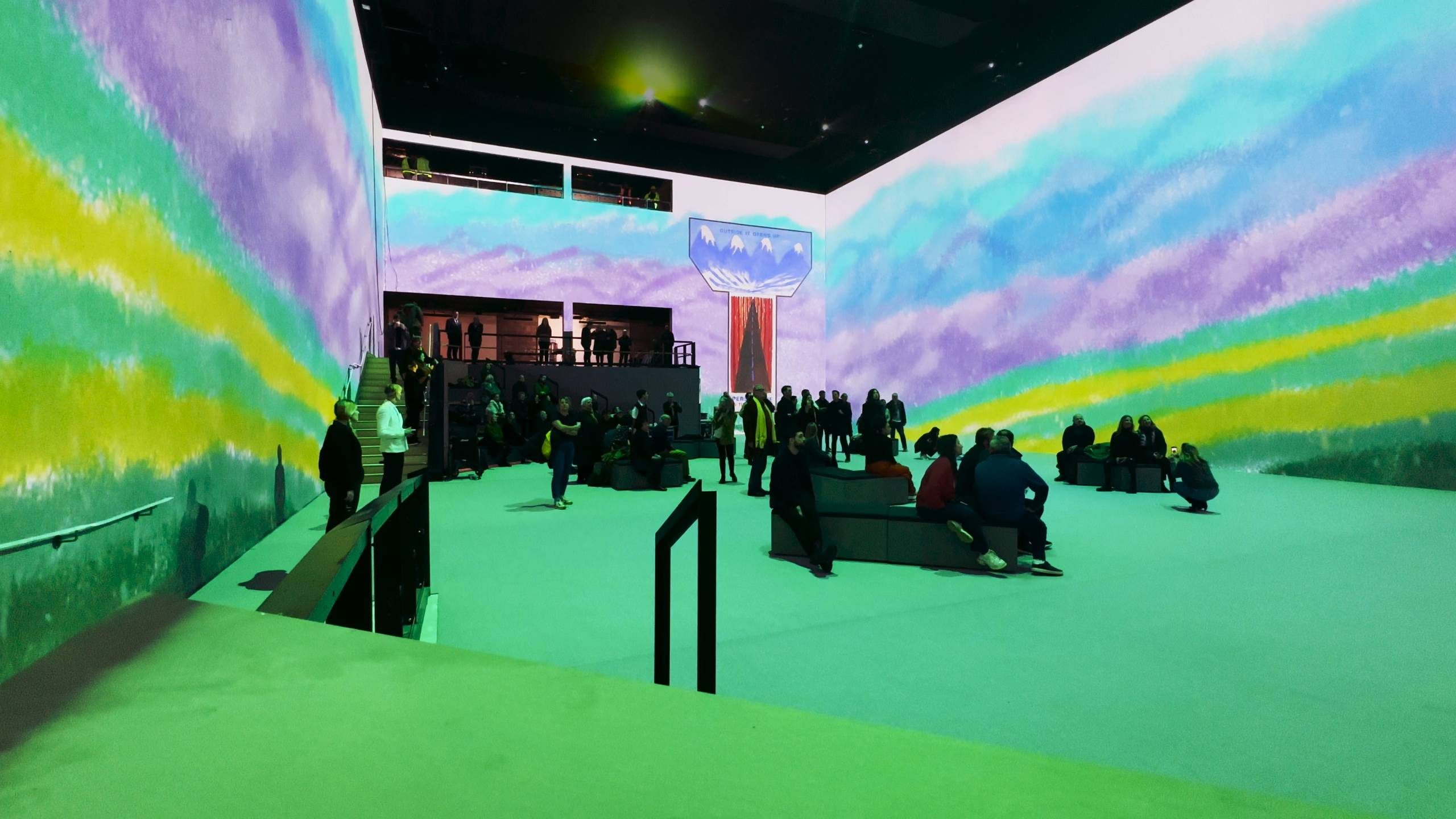 People gather in a large indoor space with colourful, immersive digital projections on the walls in shades of blue, purple, green, and yellow, creating an artistic, vibrant atmosphere. Some people are seated, others are standing.