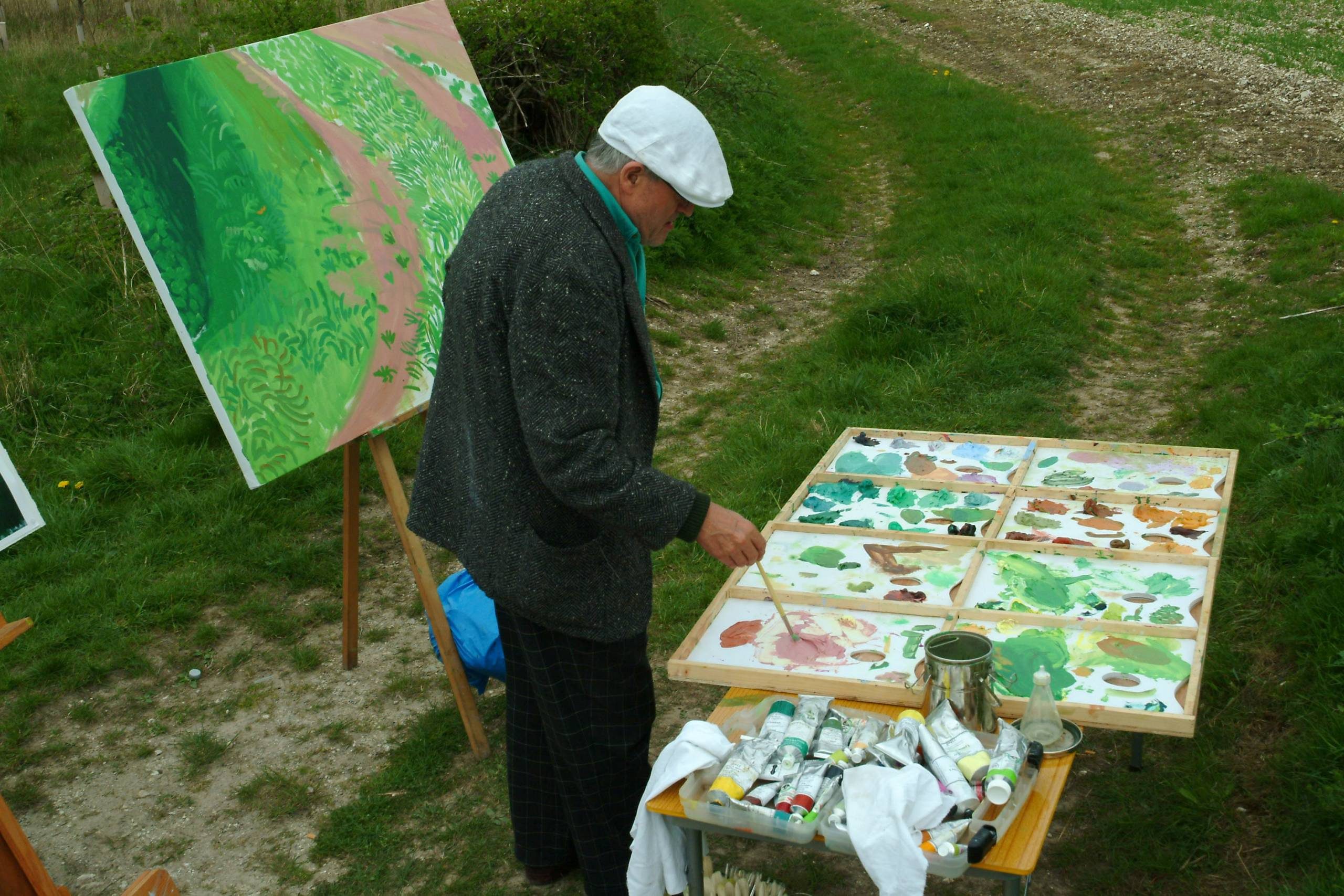 An artist wearing a white cap and dark jacket stands outdoors by a table with paints and brushes, working on a bright, leafy green landscape painting set up on an easel beside a grassy path.