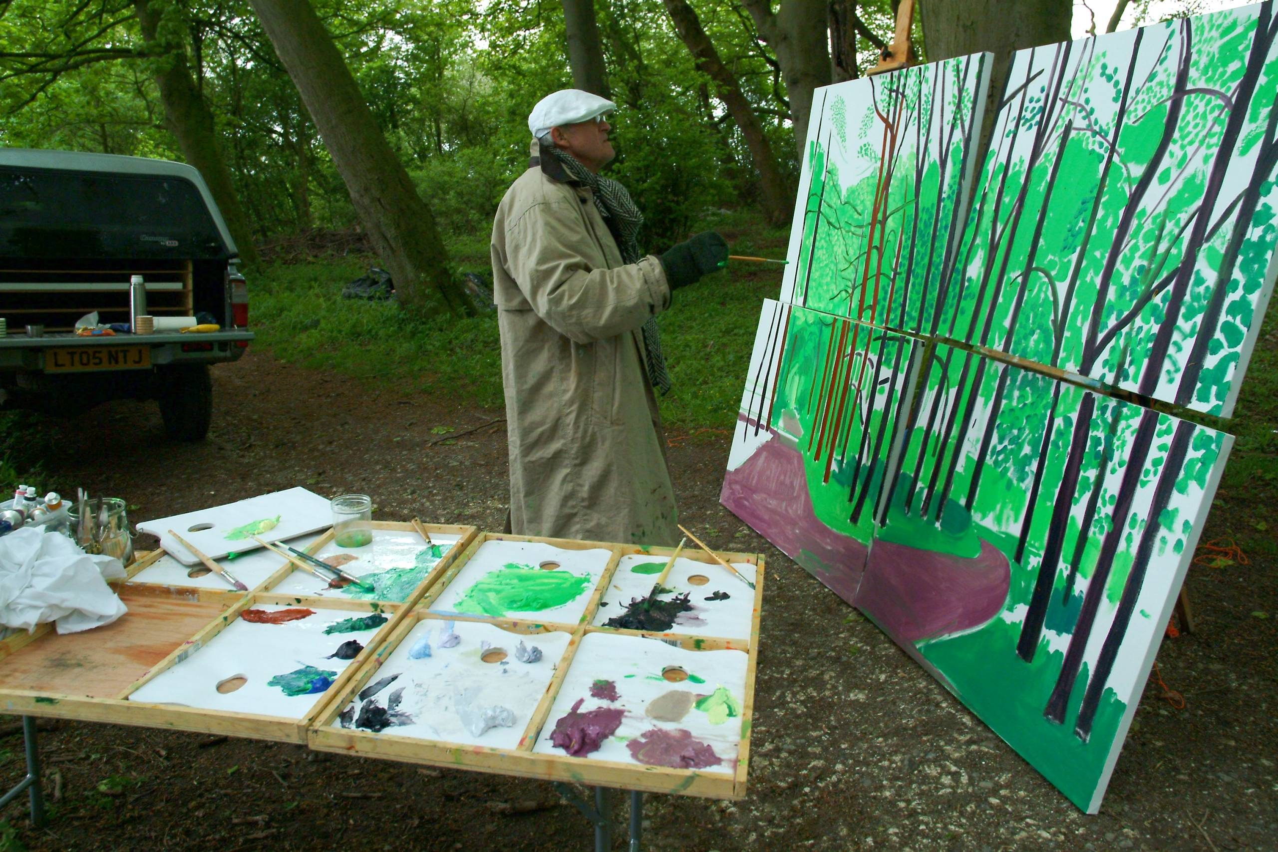 An artist wearing a long coat and white cap paints a large canvas of a green forest scene outdoors. A table nearby holds paint, brushes, and supplies, and a parked vehicle is visible in the background among tall trees.