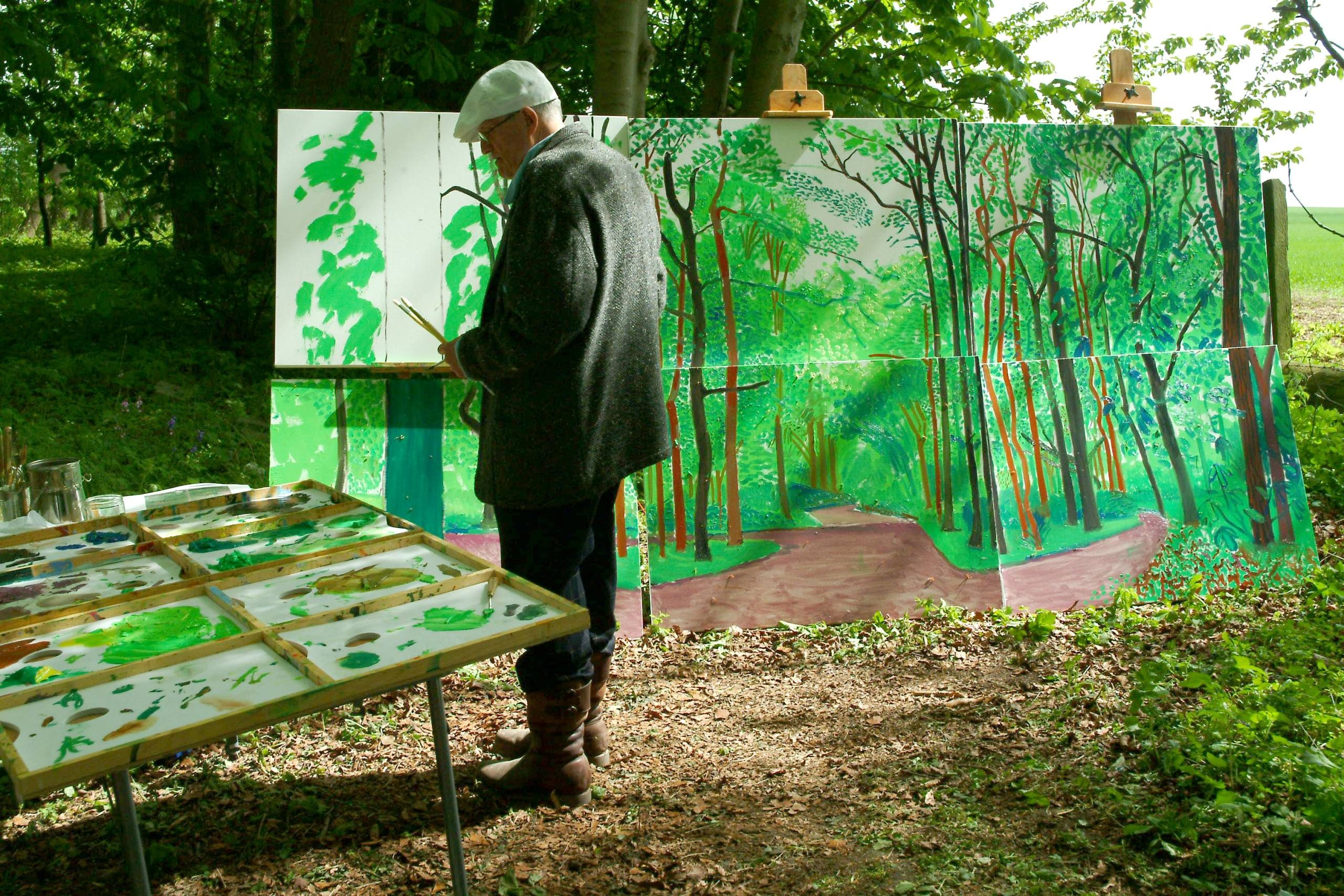 An artist stands outdoors in front of a large, colourful painting of a green forest, with paint palettes and brushes on a table nearby. Trees and sunlight surround the scene.
