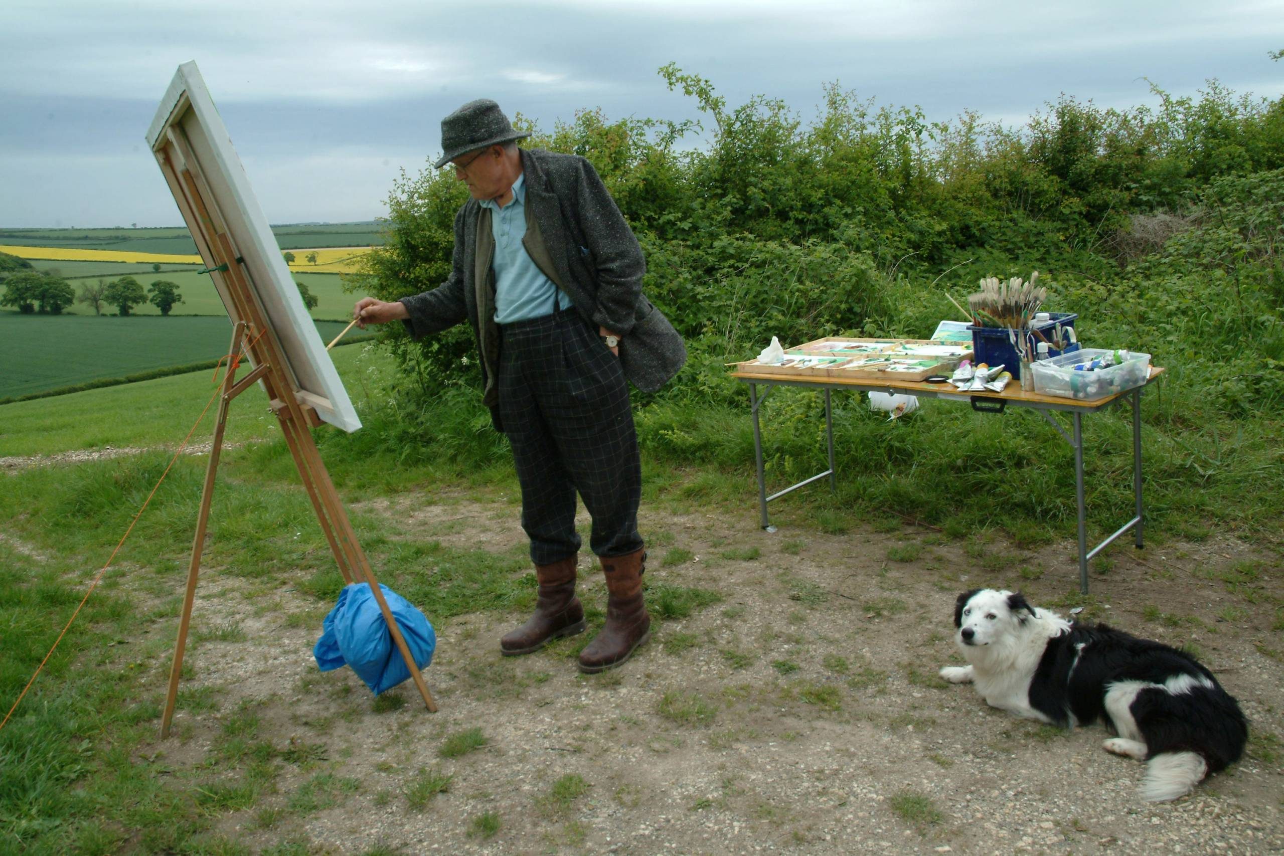 An older man in a hat and boots paints on a canvas at an easel outdoors, with art supplies on a table nearby. A black and white dog lies on the ground beside him. Fields and greenery are in the background.