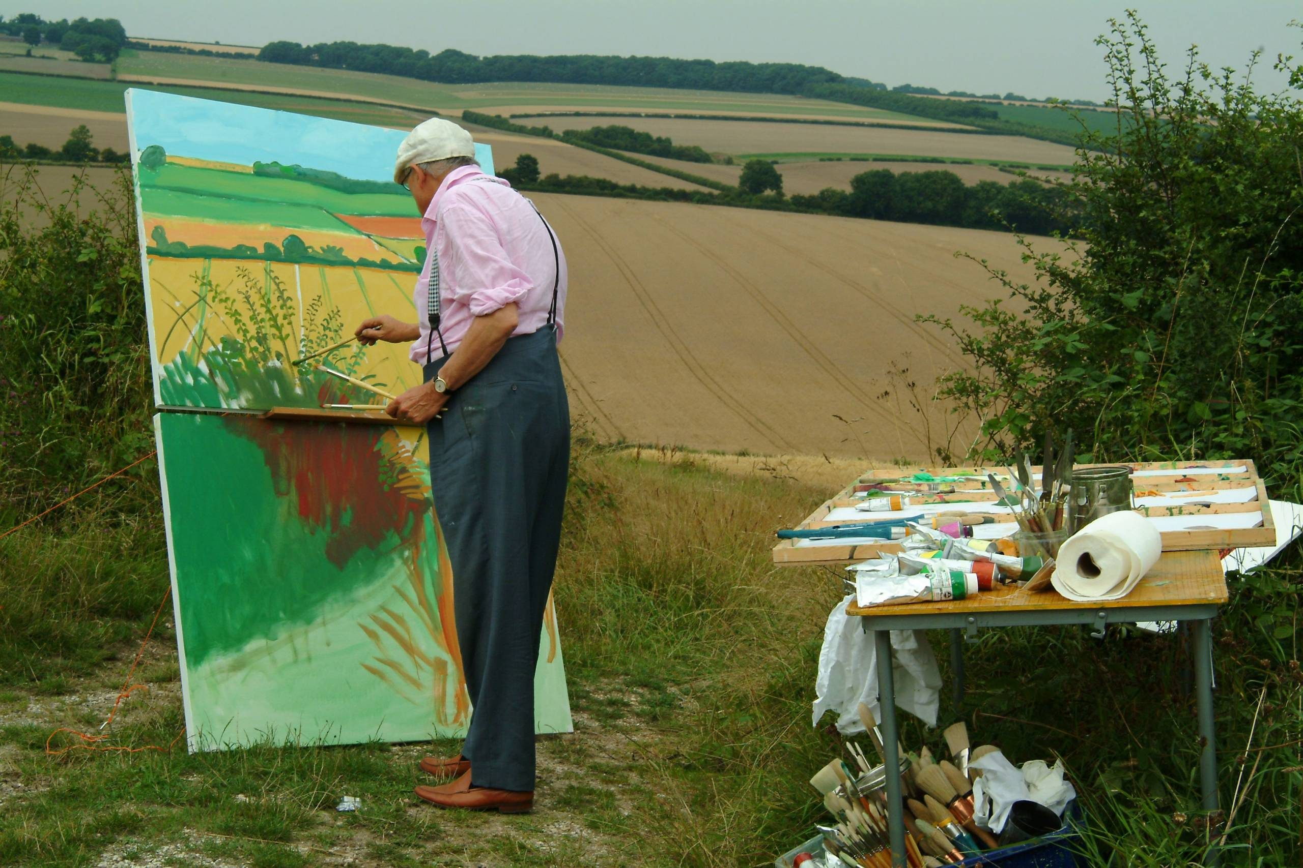 An artist in a white hat and braces paints a colourful landscape on a large canvas outdoors, with rolling farmland in the background and art supplies laid out on a nearby table.