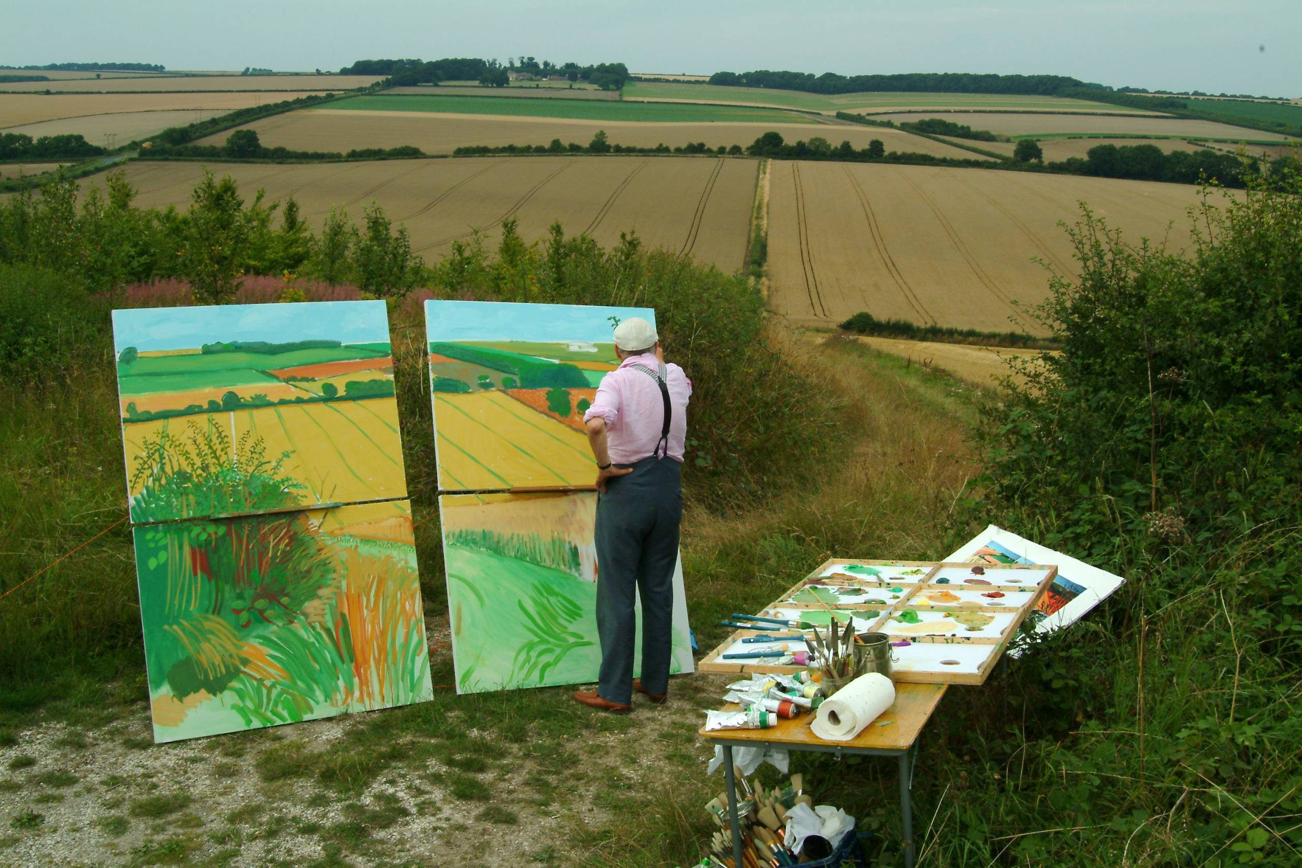 A person stands outdoors painting a landscape onto large canvases, surrounded by painting supplies on a table. The scene overlooks rolling fields and farmland under a cloudy sky.