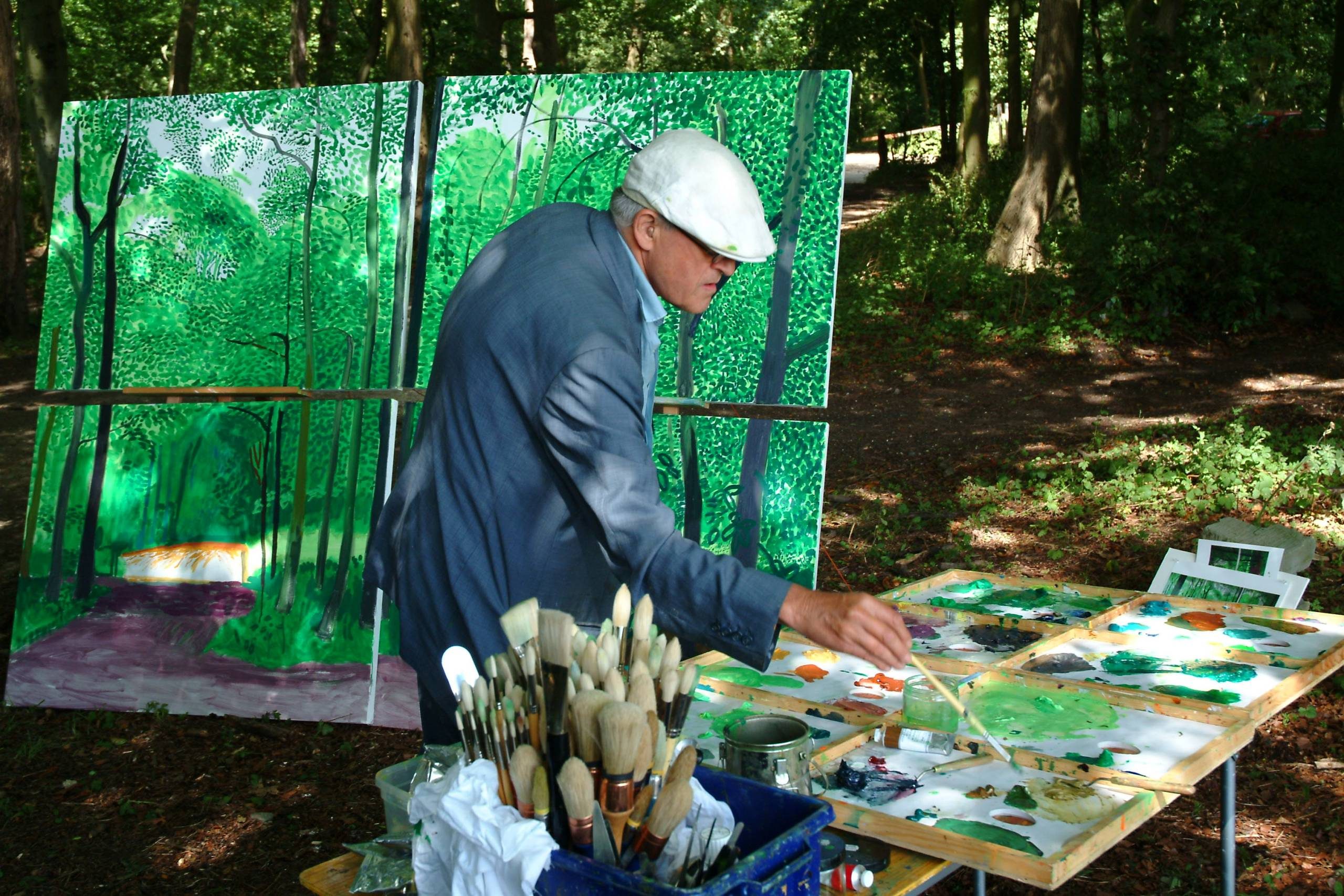 An artist in a light cap and blue jacket paints outdoors, surrounded by tall trees. He works at a table filled with brushes and paints, whilst a large green forest-themed canvas stands behind him.