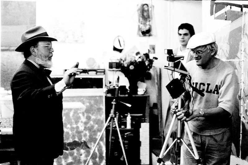 A black-and-white photo shows two older men in a studio, one in a hat gesturing, the other in a UCLA sweatshirt adjusting a camera on a tripod. A young man stands in the background. Art and equipment fill the room.