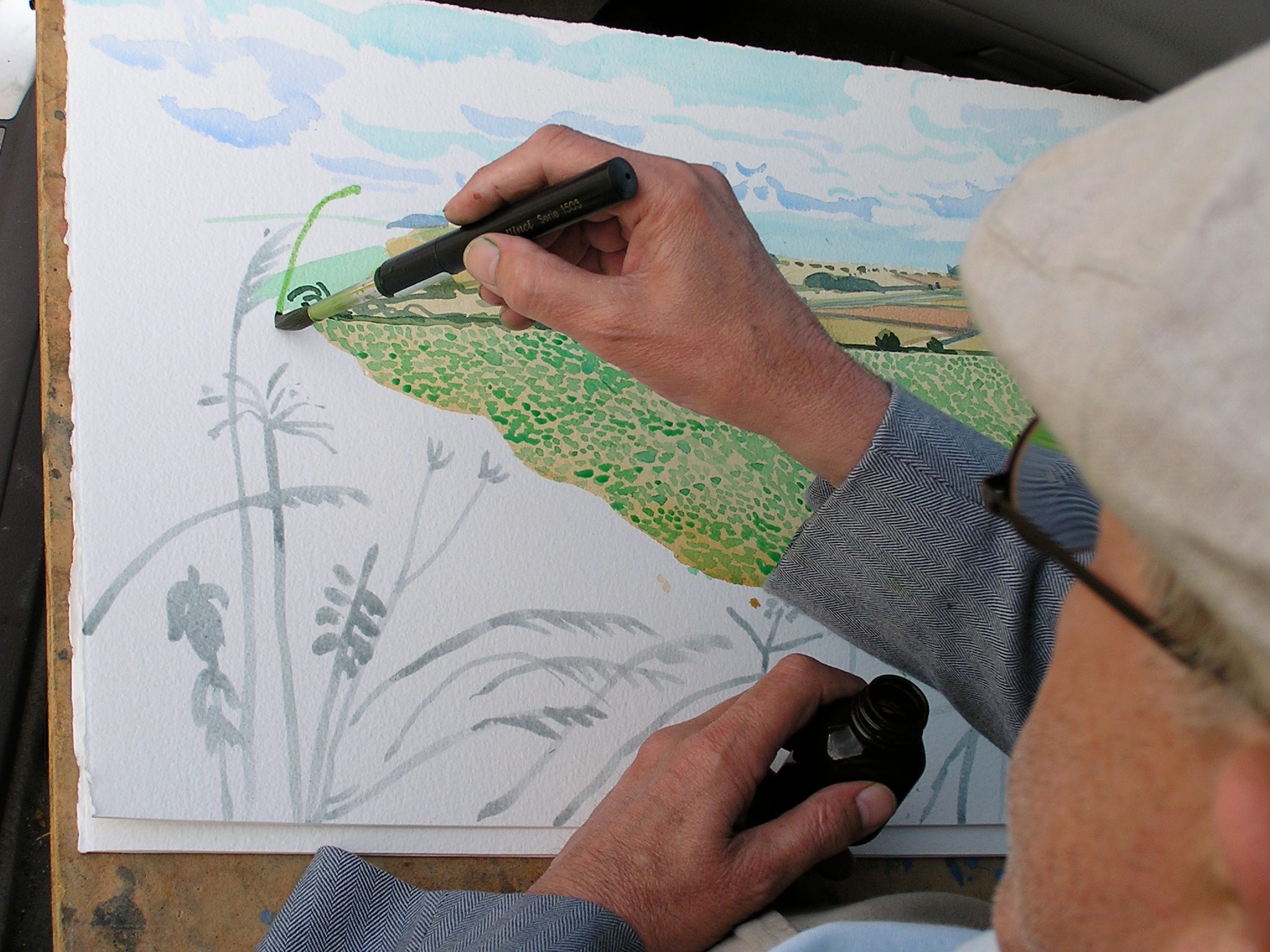 A person wearing a light cap and blue jacket is drawing a countryside landscape on paper with felt-tip pens, detailing green fields and plants under a blue sky.