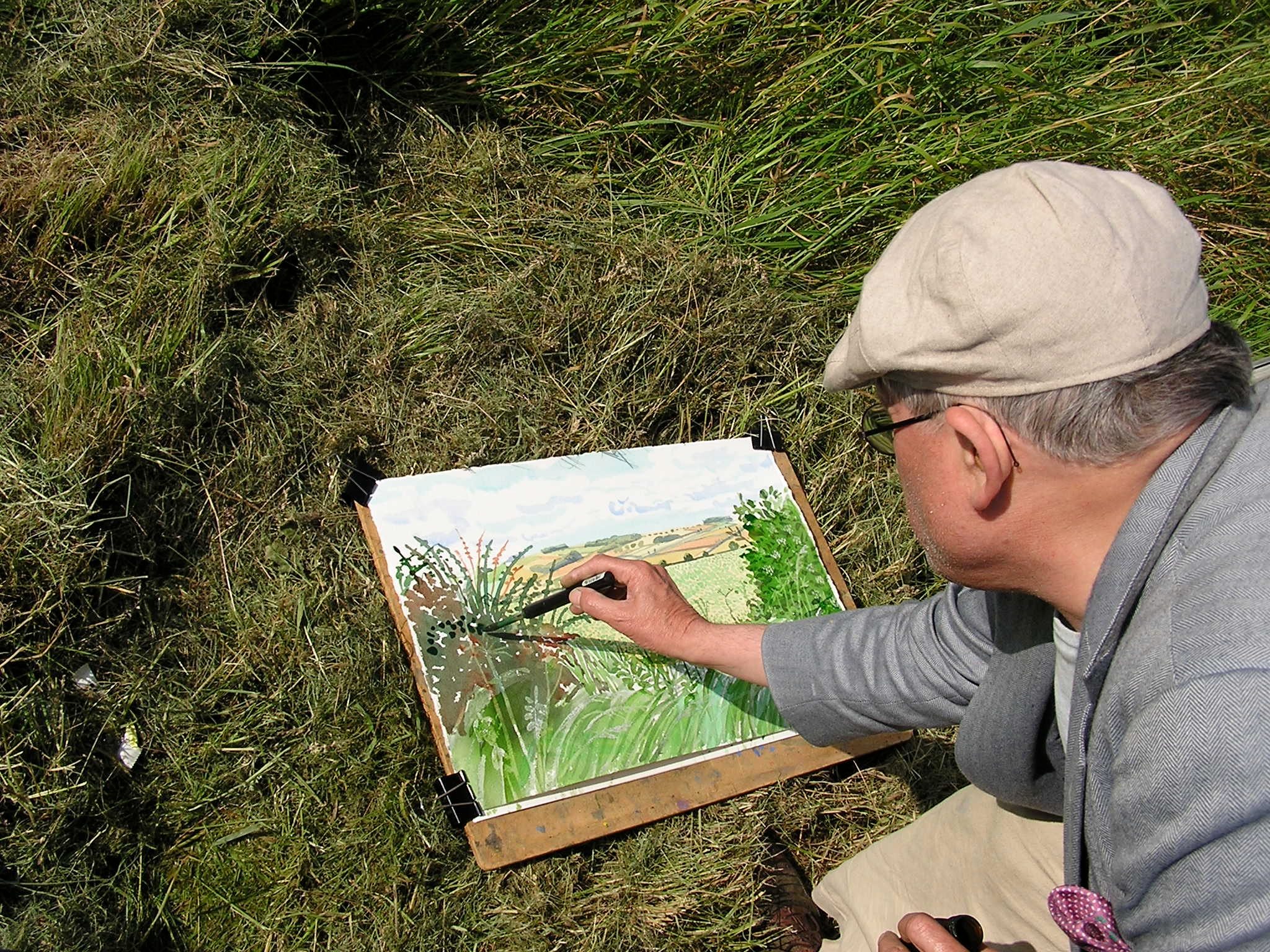 A person wearing a cap and glasses is sitting outdoors on grass, painting a landscape scene with green plants and fields on a piece of paper using a brush. The painting is clipped to a board.