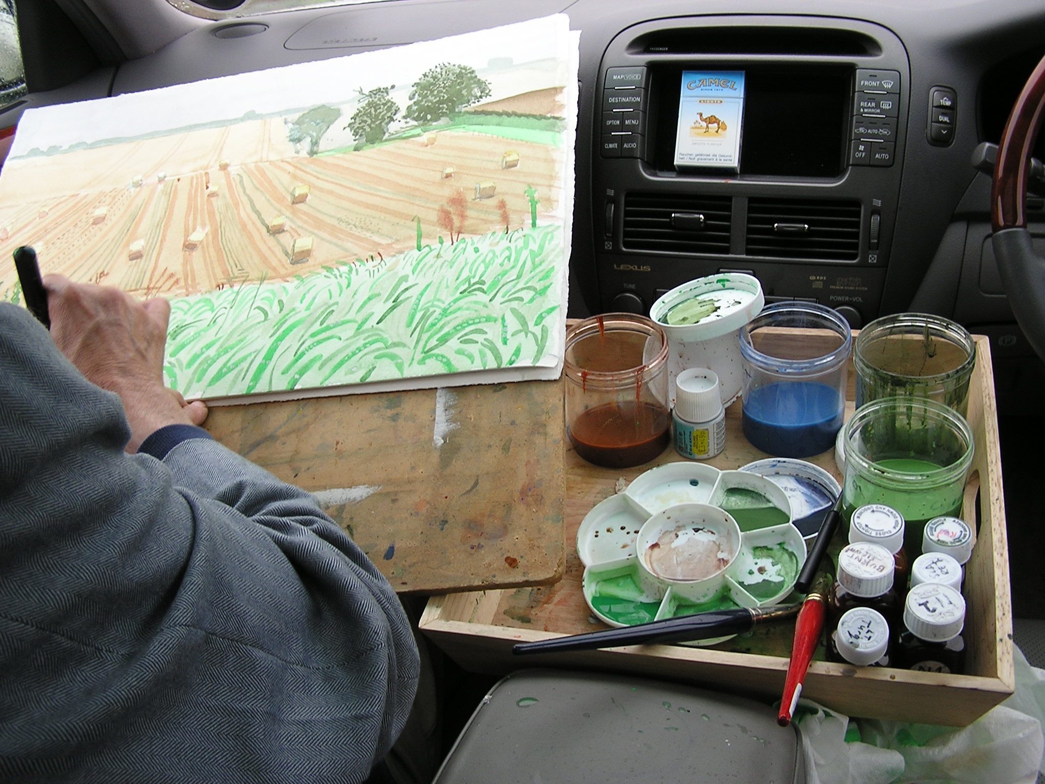 A person paints a rural landscape scene from inside a car, with paint jars, brushes, and palettes arranged on a tray on the passenger seat. The painting shows fields, hay bales, and trees.