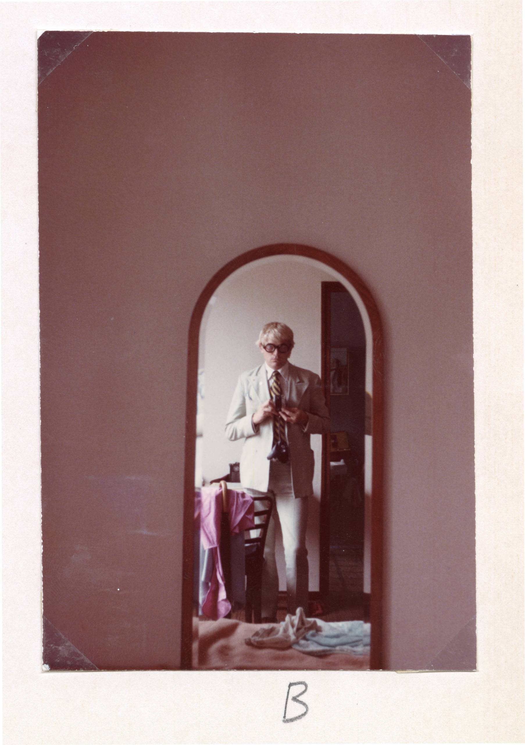 A person wearing glasses and a light-coloured suit is taking a photo of themselves in a tall, arched mirror. The background includes a bed with clothes on it. The photo is faded and has the letter B written below it.