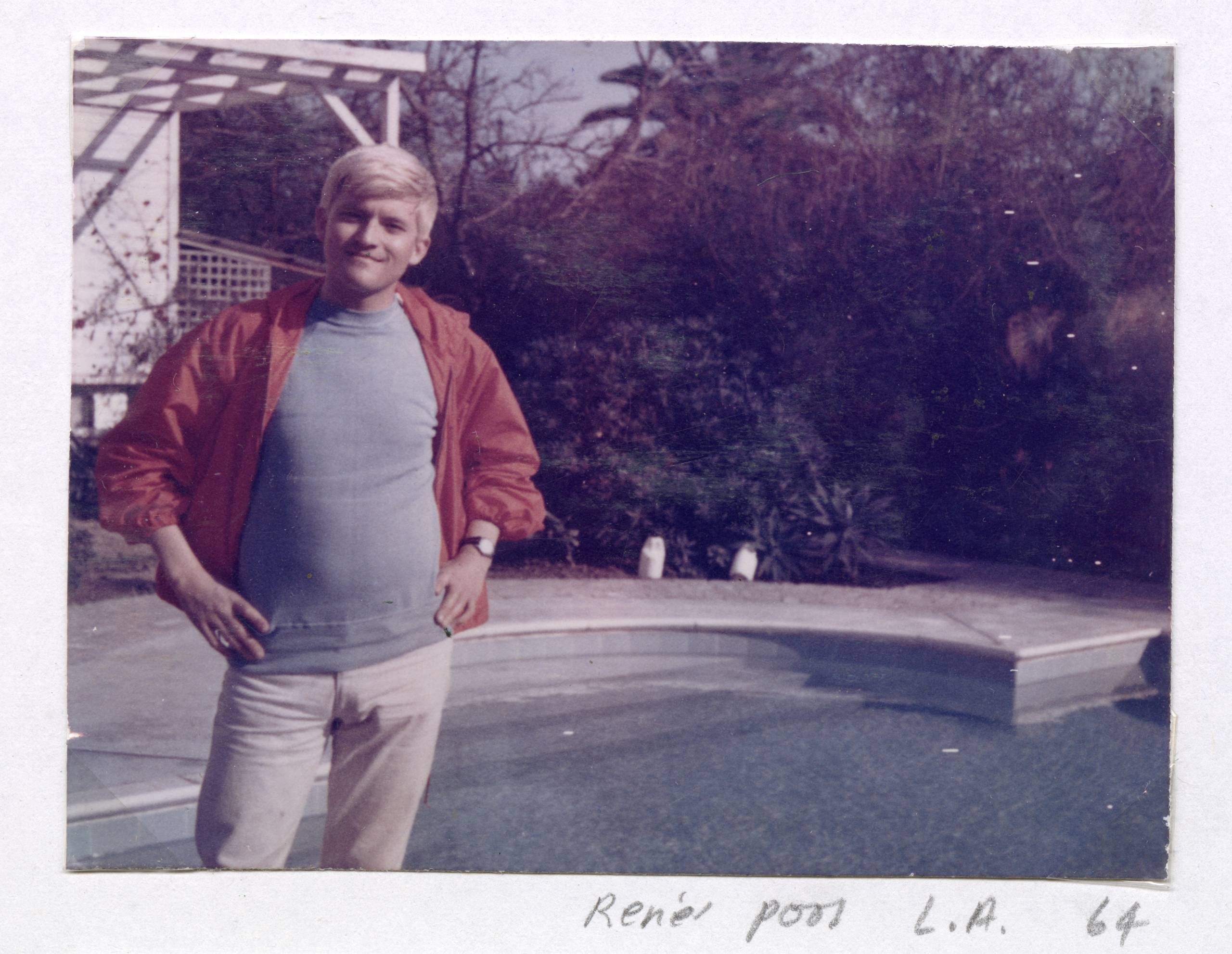 A young man with blond hair, wearing a red jacket and light trousers, stands smiling by a swimming pool. Trees and bushes are in the background. The handwritten caption reads “René pool L.A. 64”.
