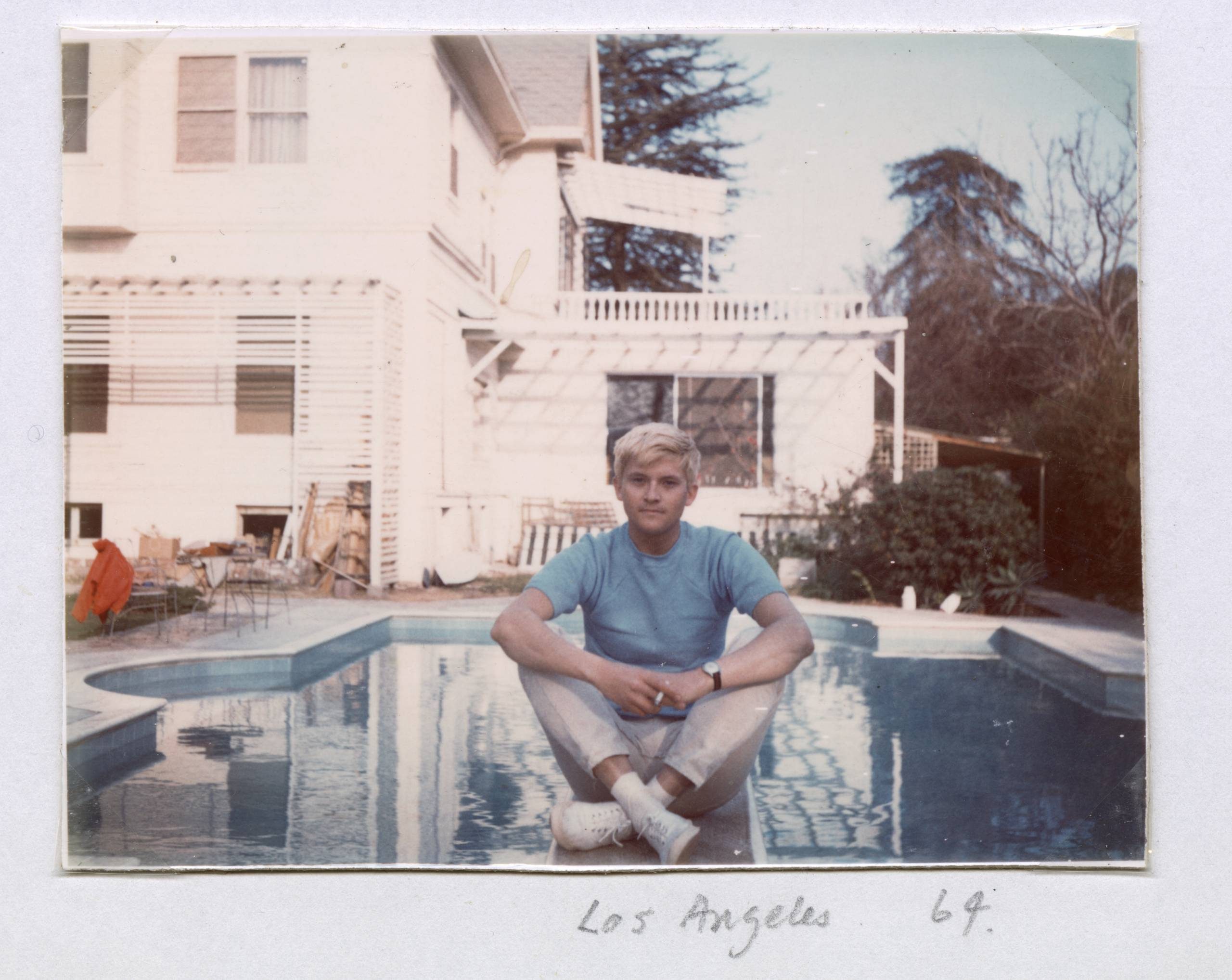 A young man in a blue T-shirt and light trousers sits cross-legged at the edge of a swimming pool in front of a white house with garden furniture. Trees and shrubs are visible in the background. Los Angeles 69 is handwritten below.