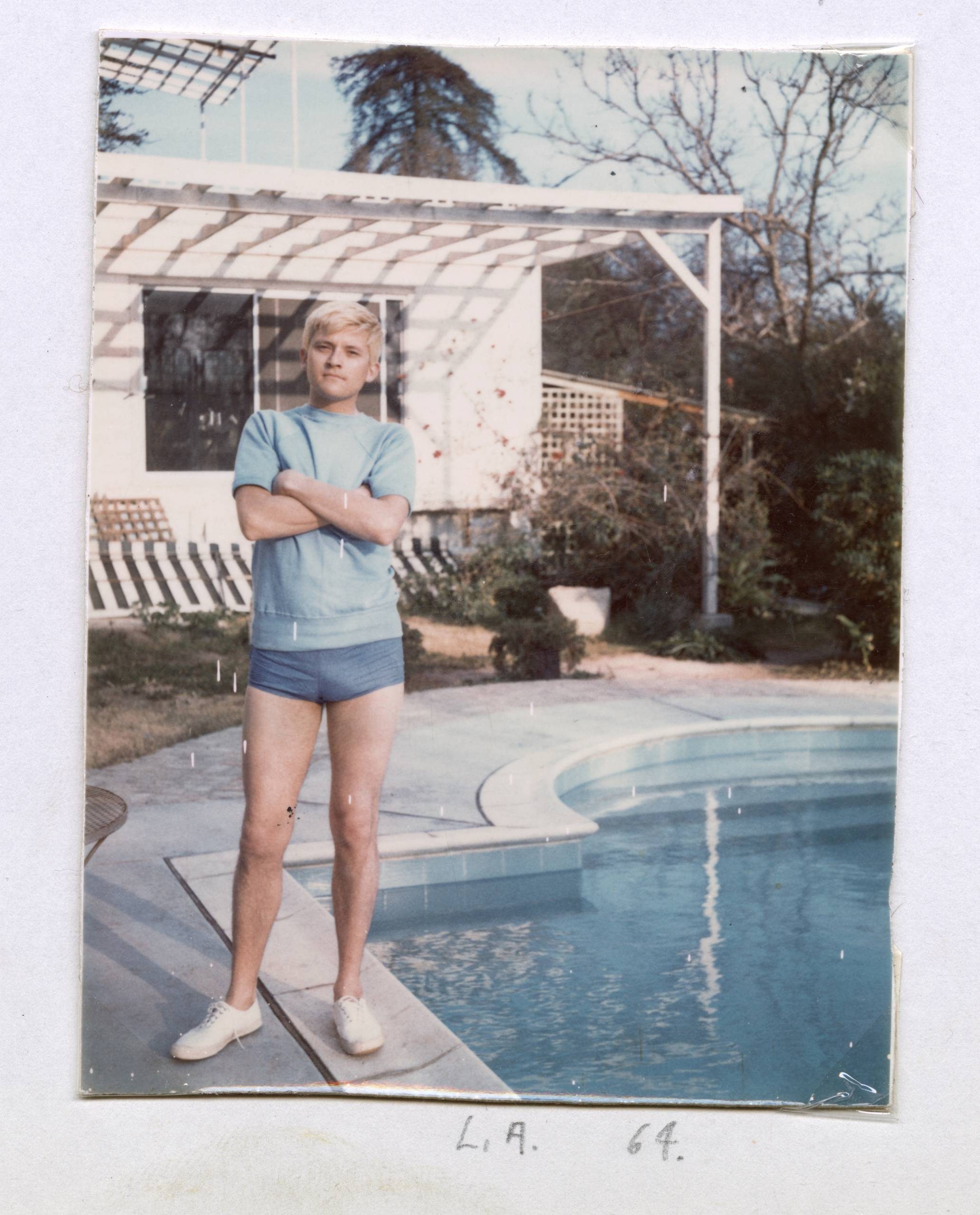A person with short blond hair, wearing a blue T-shirt and shorts, stands with arms crossed by a back garden pool. Behind them is a white house with a pergola. Handwritten text below reads L.A. 64.