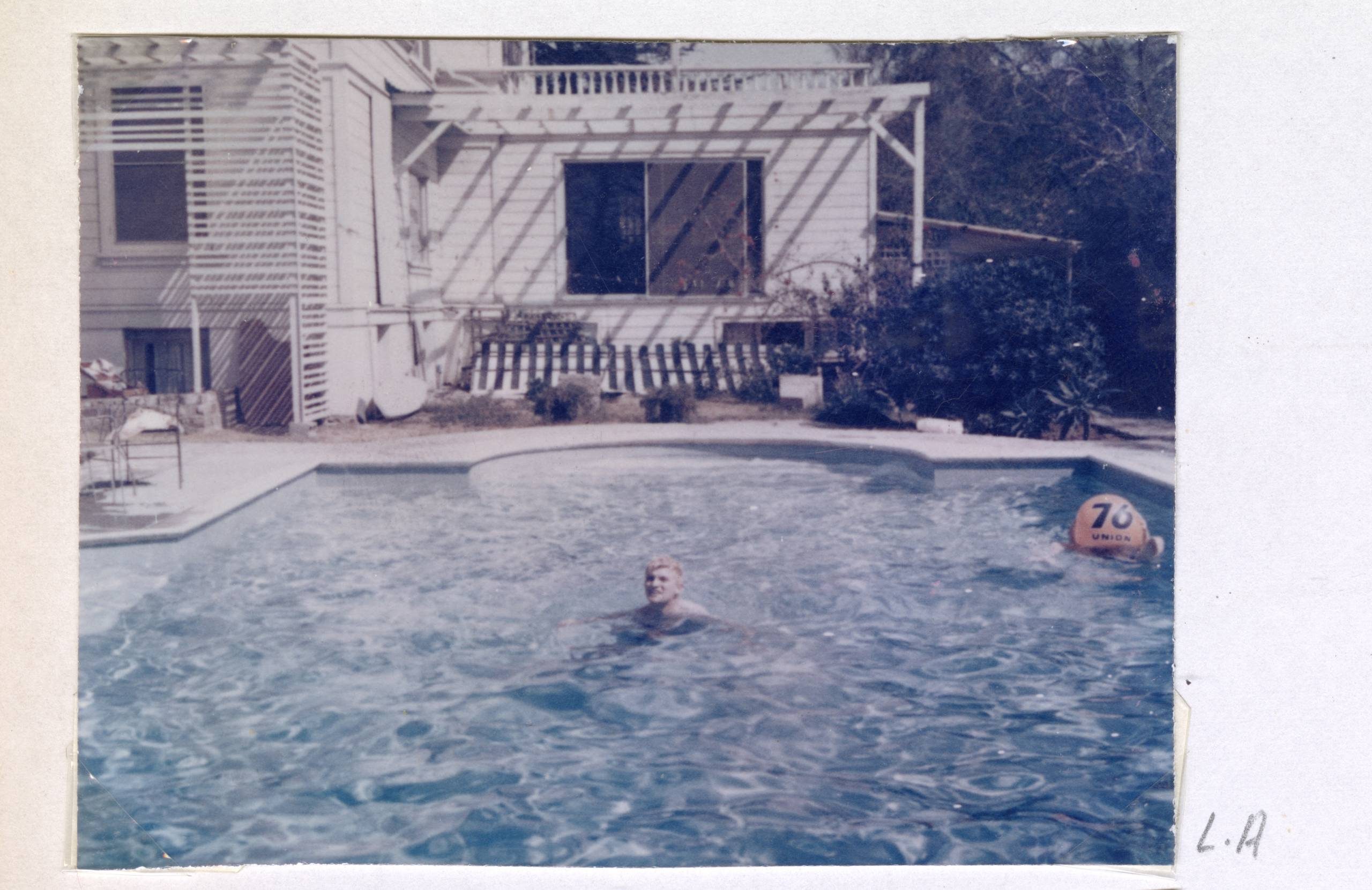 A person is swimming alone in an outdoor pool in front of a white house with a balcony and trellis. A buoy marked 70 floats nearby. The word L.A. is written on the photo’s border.