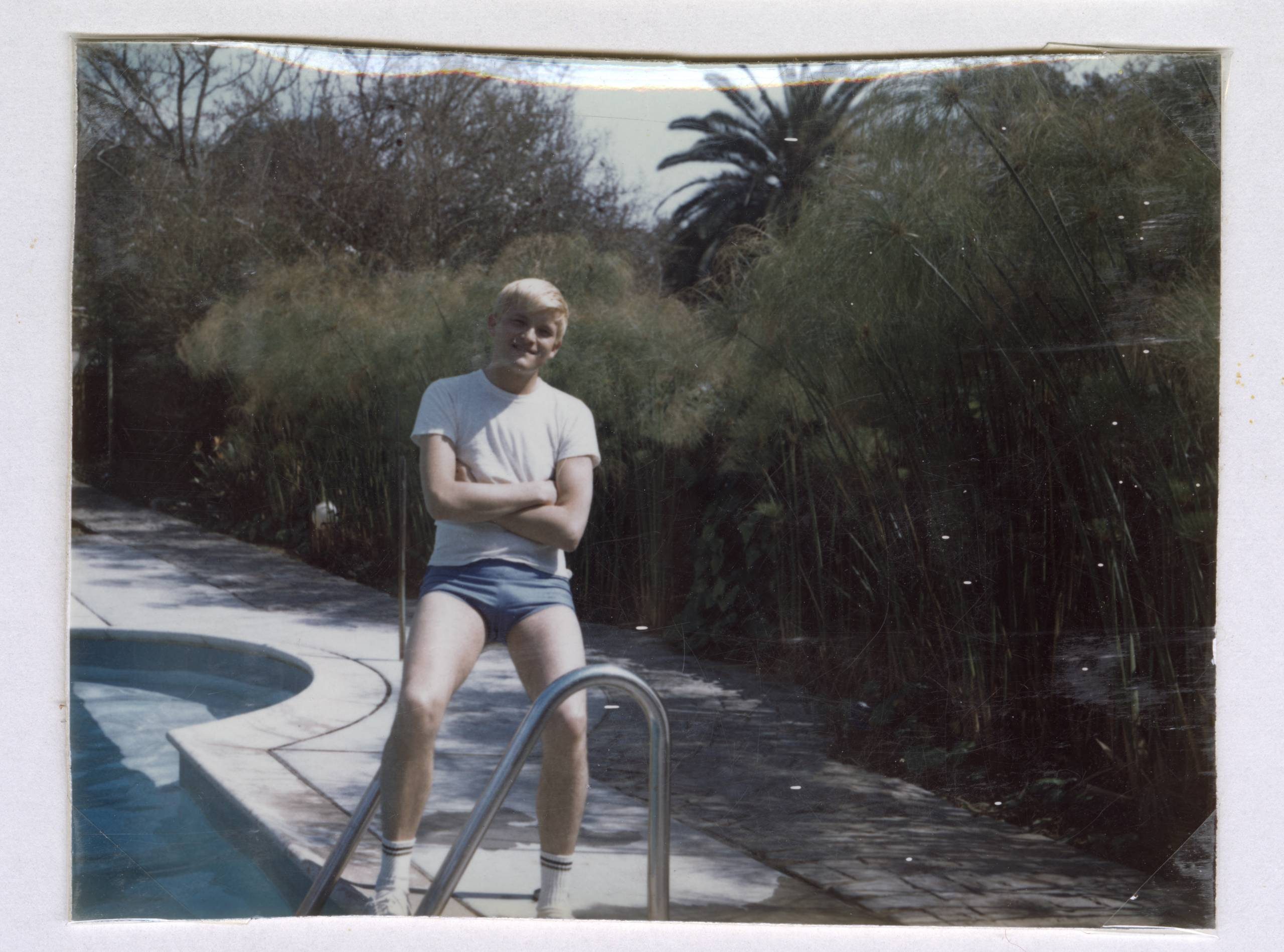A young person with short blond hair, wearing a white T-shirt and blue shorts, stands with arms crossed by the metal railing of a back garden pool, surrounded by greenery and palm trees.