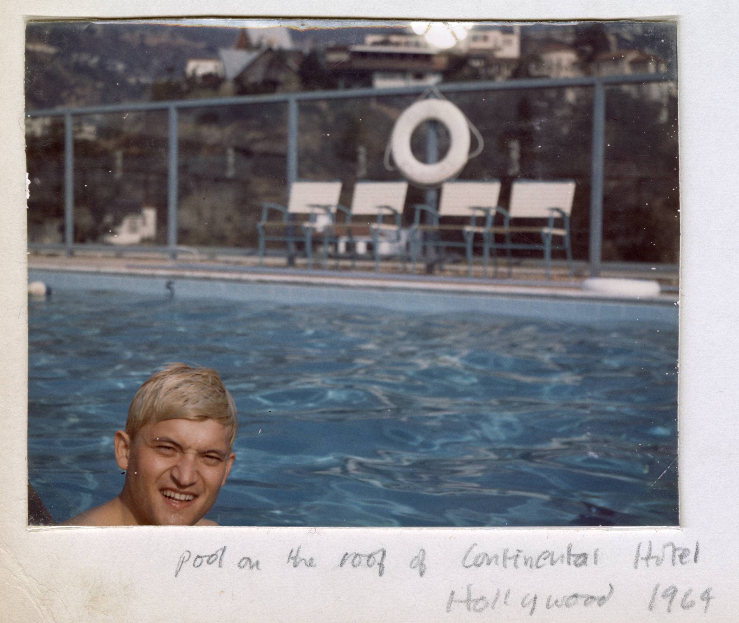 A person with short blond hair smiles in a rooftop swimming pool. Behind them are pool chairs, a life preserver, and a glass fence, with houses and hills in the background. Handwritten text reads: pool on the roof of Continental Hotel Hollywood 1964.