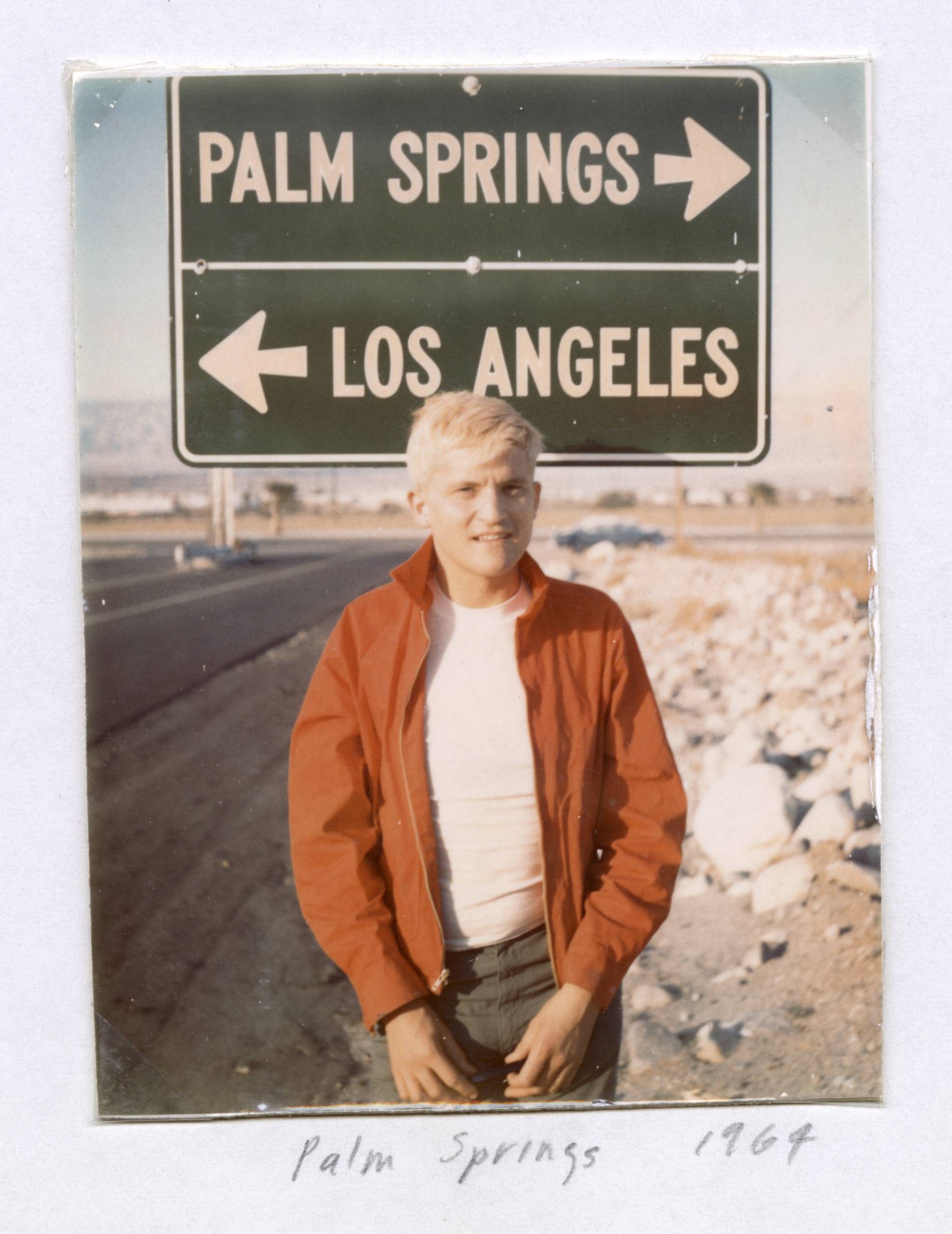 A young man with short blond hair and a red jacket stands on a roadside near a sign pointing to Palm Springs and Los Angeles. The handwritten caption below reads “Palm Springs 1967”.