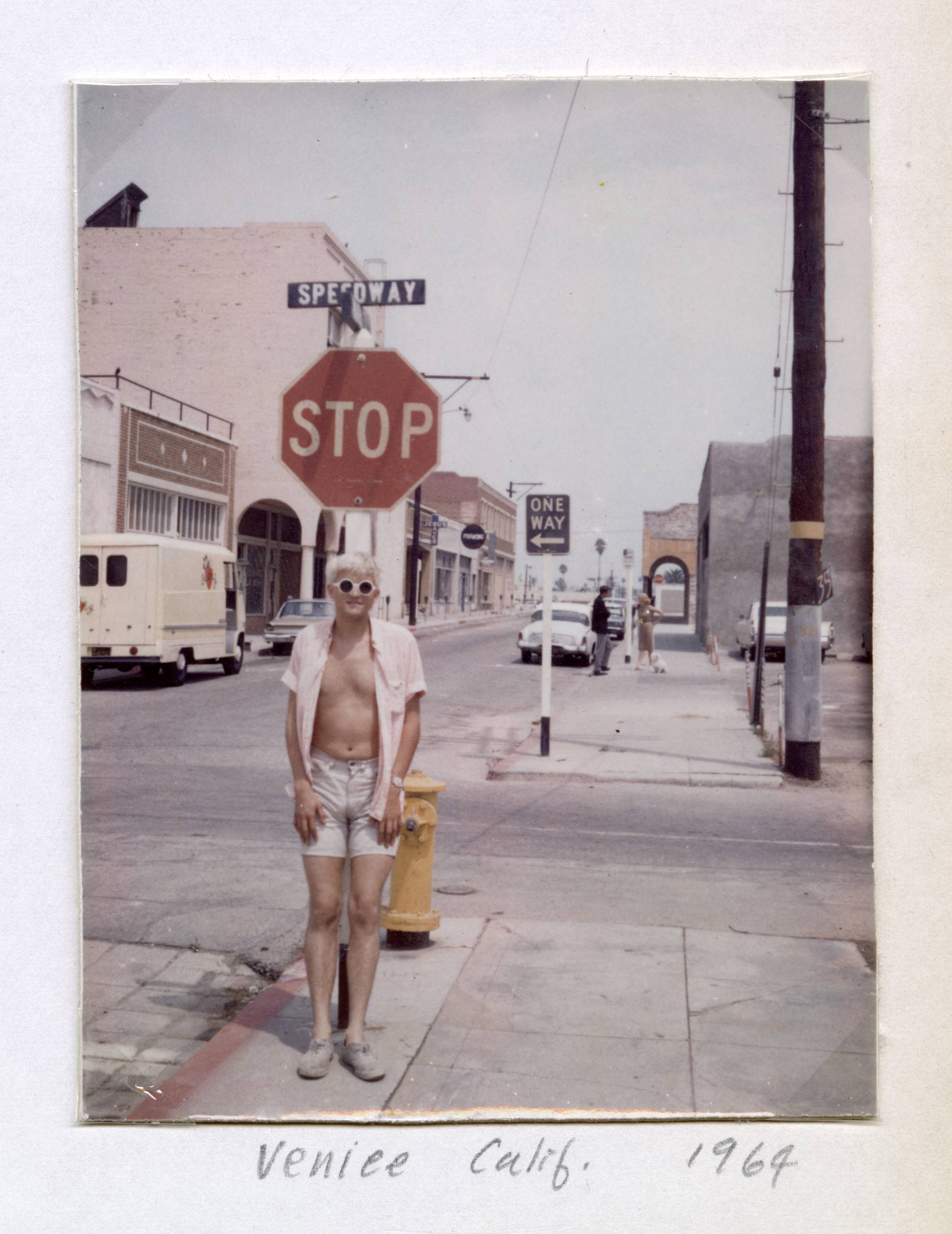 A person in shorts and sunglasses stands by a yellow fire hydrant in front of a stop sign on a street in Venice, California. The photo, labelled Venice Calif., 1964, shows buildings and a van in the background.