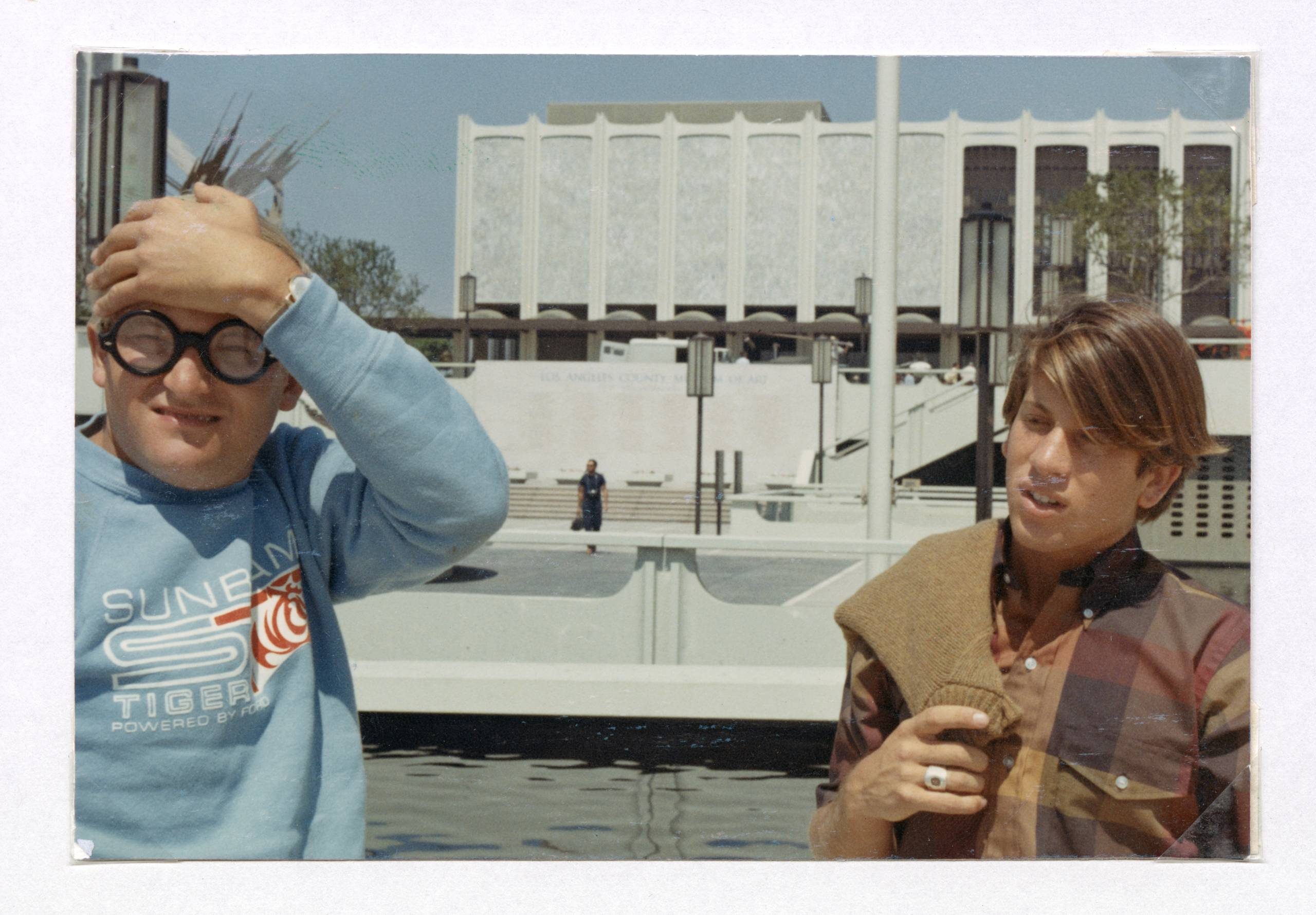 Two boys stand in front of a modern white building. One wears thick glasses and a blue jumper, holding his head and smiling. The other, in a striped shirt with a jumper over his shoulder, looks away thoughtfully.