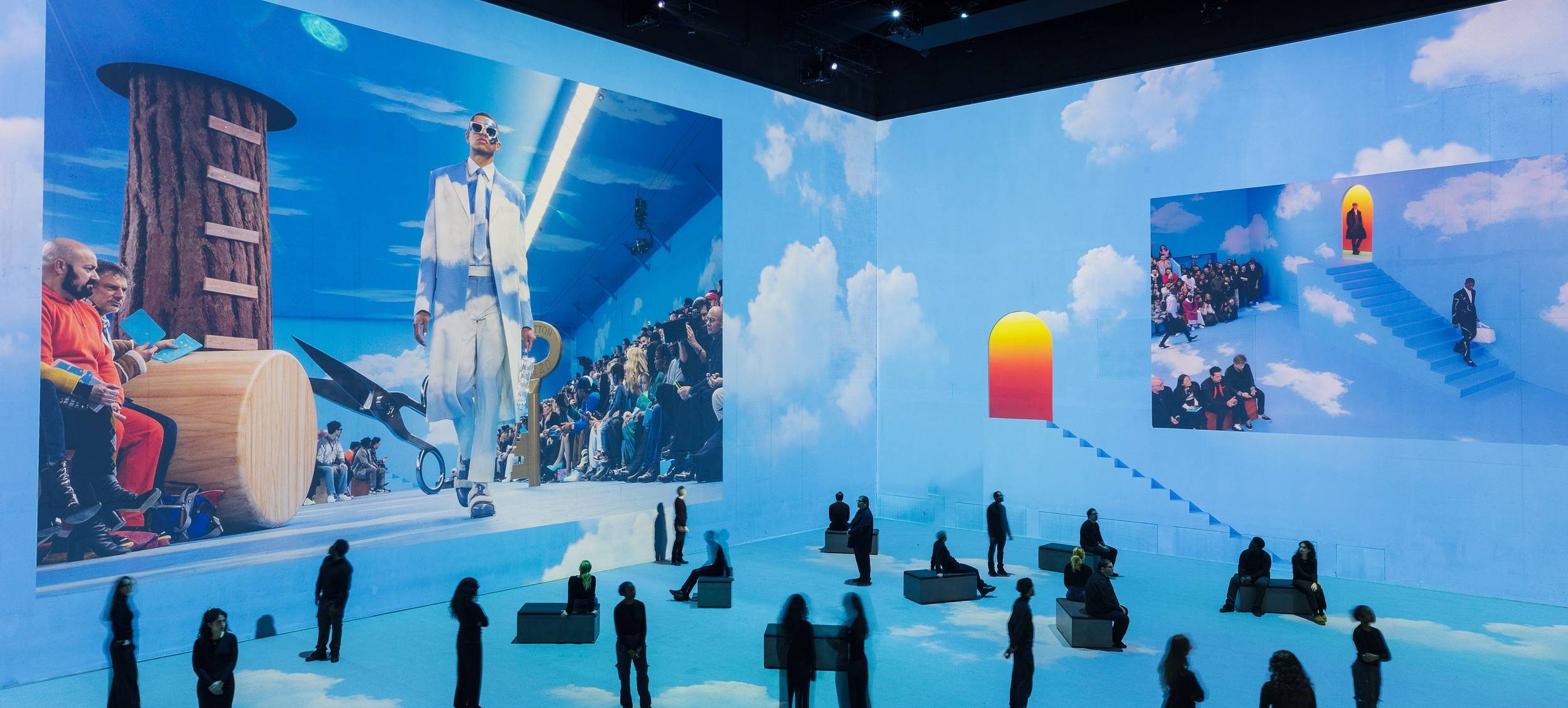 People stand in a large room with sky-blue walls and floor, covered in cloud patterns. The walls display oversized images of a fashion show with models walking along a catwalk and audiences seated nearby.