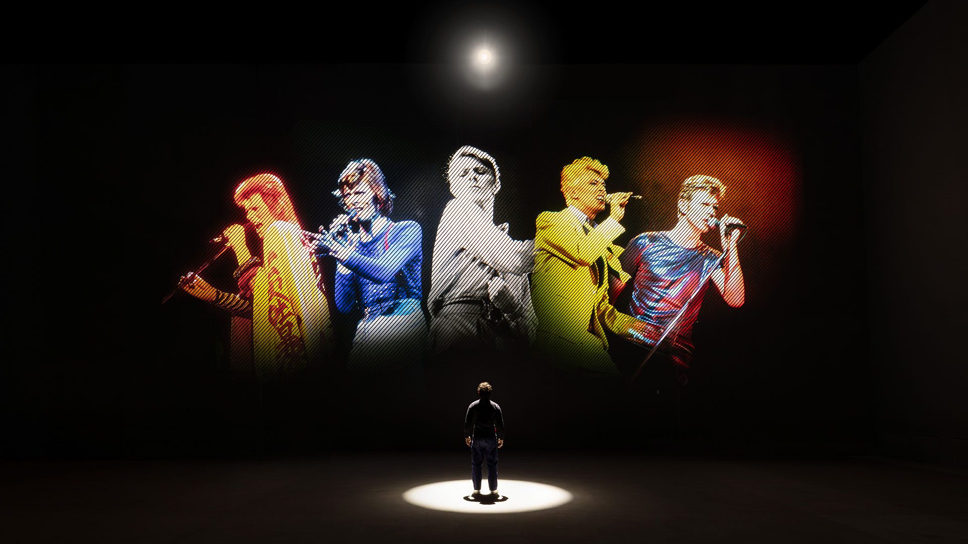 A person stands in a spotlight, gazing at a large, colourful projection of five different singers performing on stage, each illuminated in red, yellow, white, gold, and blue lighting.