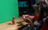 A person adjusts a clay figure on a desk in front of a green screen; a computer in the background displays the figure on the screen.