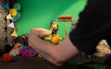 A person adjusts a claymation dog figure holding a broom on a miniature set, with a green screen and colourful decorations in the background.