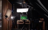A backstage studio set-up with lights, a camera on a tripod, and a small green screen on a table. Props and equipment are arranged around the workspace, suggesting a filming or animation project in progress.
