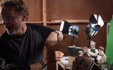 A man works on a stop-motion set with Wallace and Gromit models, surrounded by lights and camera equipment, in a studio setting.
