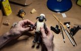 A person holds a clay model of a sheep on a worktable surrounded by sculpting tools, art supplies, and other model parts, suggesting a stop-motion animation setting.