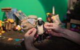 Hands hold a claymation sheep figure in a small, detailed set with colourful miniature props, bunting, and a bright green backdrop.