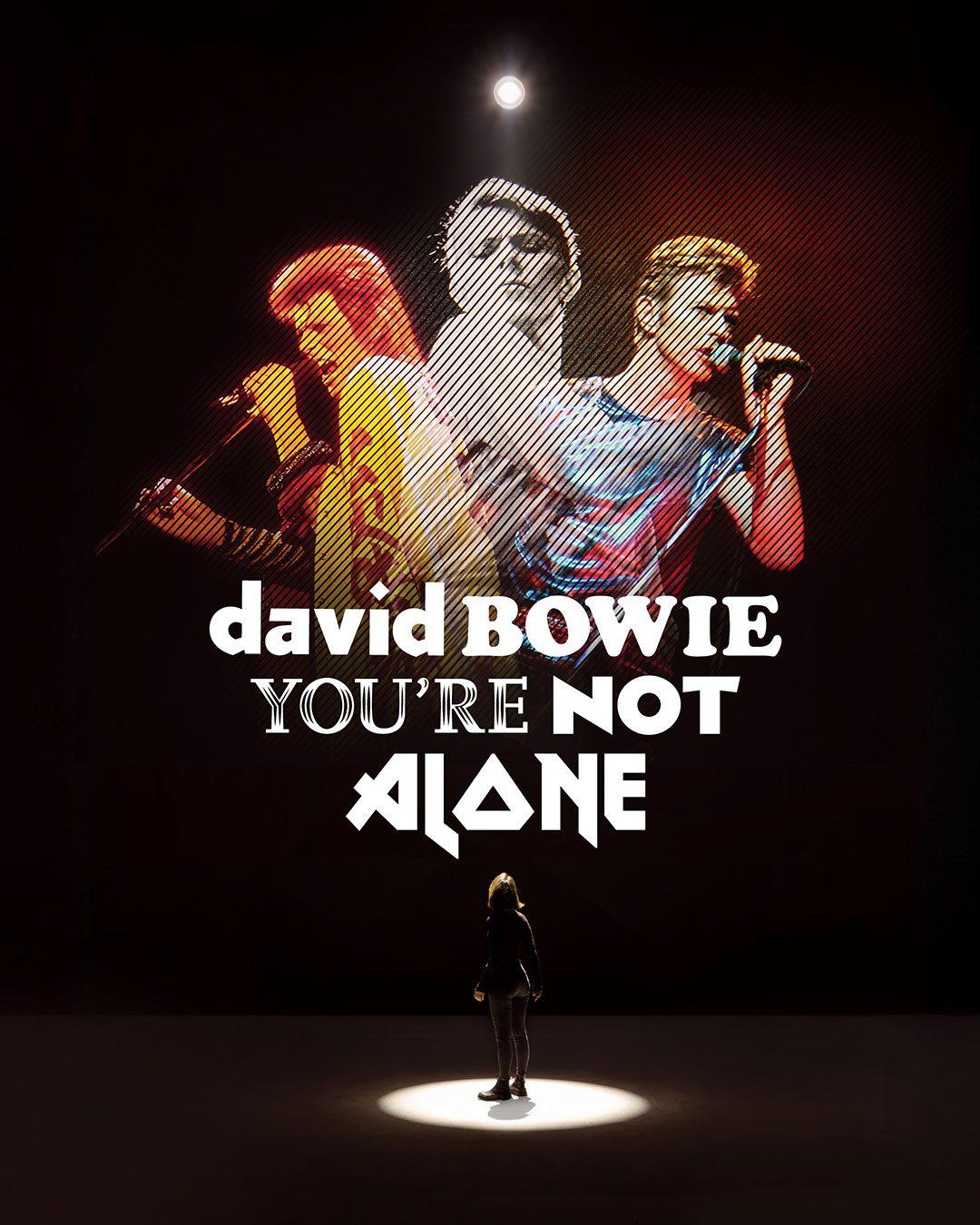 A person stands in a spotlight, gazing up at three images of David Bowie performing, with the text “david BOWIE YOU’RE NOT ALONE” displayed in bold white letters.