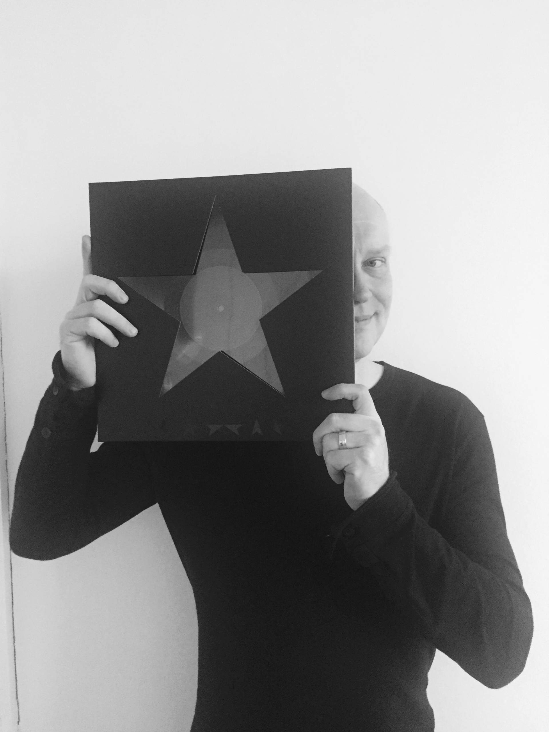 A person in a black shirt holds a square album cover with a large star cut-out, partially covering their face, whilst smiling in front of a plain white wall.