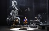 Three musicians perform on stage with electronic equipment and a cello, as a large black-and-white photo of a cellist is projected behind them.