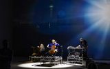 Three musicians perform on a dimly lit stage with a cello, keyboard, and electronic equipment. A cityscape and tower are projected on the wall behind them, illuminated by a bright spotlight.