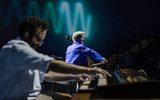 A man in focus plays a cello on stage, wearing a blue shirt, whilst another man in the foreground plays a keyboard. Blue light waves are projected onto the dark background.
