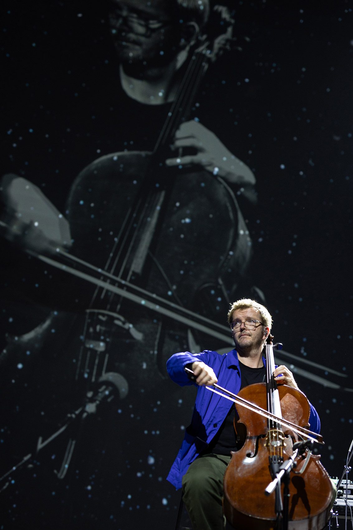 A cellist in a blue shirt plays on stage, with a black-and-white projection of himself playing the cello displayed behind him.