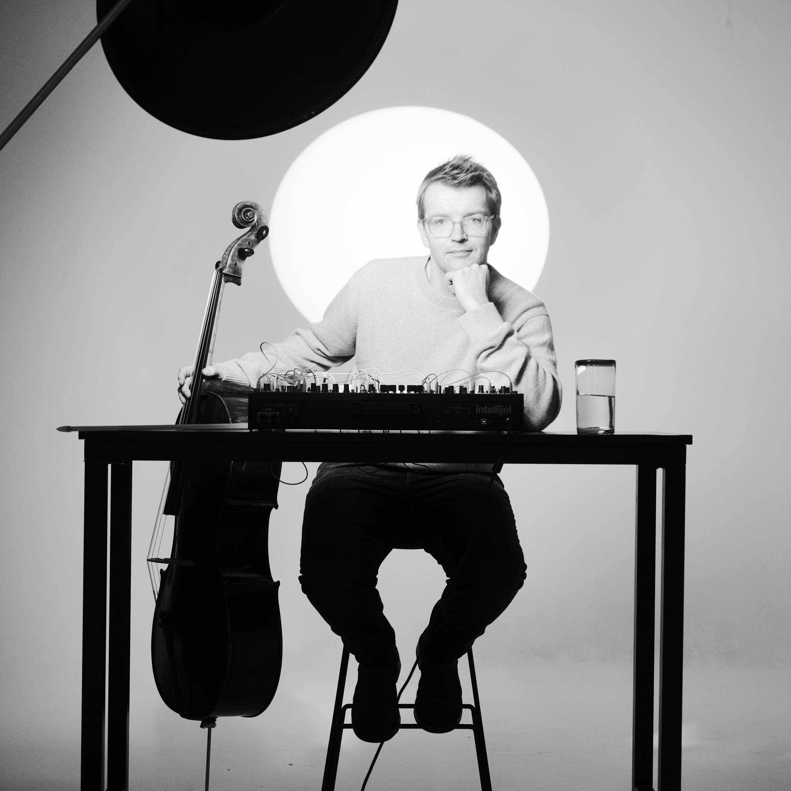 A person sits on a stool at a table with a cello, audio equipment, and a glass of water. A bright circular light is behind them, creating a dramatic black and white scene.