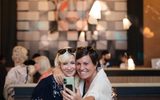 Two smiling women take a selfie together in a lively café, with a large, colourful mural of a face in the background and other people chatting around them.