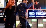 A woman in a black dress and a man in a blue star-patterned jacket dance together in front of a DJ booth with a sign reading Tom Hackers Mobile Discotheque. The DJ is playing music in the background.