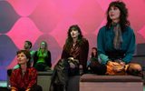 Three people sit on benches in a modern, colourful room with a pink and purple geometric wall. They appear to be watching something, whilst a few others sit and observe in the background.