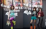 Three people in colourful, retro outfits pose in front of a mural of David Bowie. One, in a green dress and yellow boots, is energetic; the other two wear star-patterned tights and platform boots, with hands on hips.