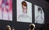 Two people sit on stage in conversation, silhouetted against large projected images of a person in iconic and artistic poses, with bold make-up and striking hair, displayed on a wall behind them.