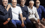 Five older men sit closely together, smiling, against a dark background. They wear casual and semi-formal outfits, including black, white, and navy blue shirts, and one wears a white suit with glasses.