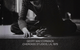 Two people sit on stage in front of a large black-and-white photo showing a person writing, with the text Geoff MacCormack Cherokee Studios, LA, 1975 displayed under the image.