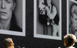 Two people stand in front of a wall displaying large black-and-white portraits of a man in different poses. One person holds a microphone and gestures whilst the other faces the photos.