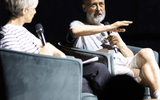 Two people sit in armchairs on stage, holding microphones and engaging in conversation. One person gestures mid-sentence whilst the other listens attentively. The background is dark, focusing attention on the speakers.