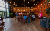 A spacious, modern lounge with wooden floors, hanging lights, and a curved bar at the centre. People are seated at the bar, and a Spotify logo is visible on an orange wall decorated with framed photos.